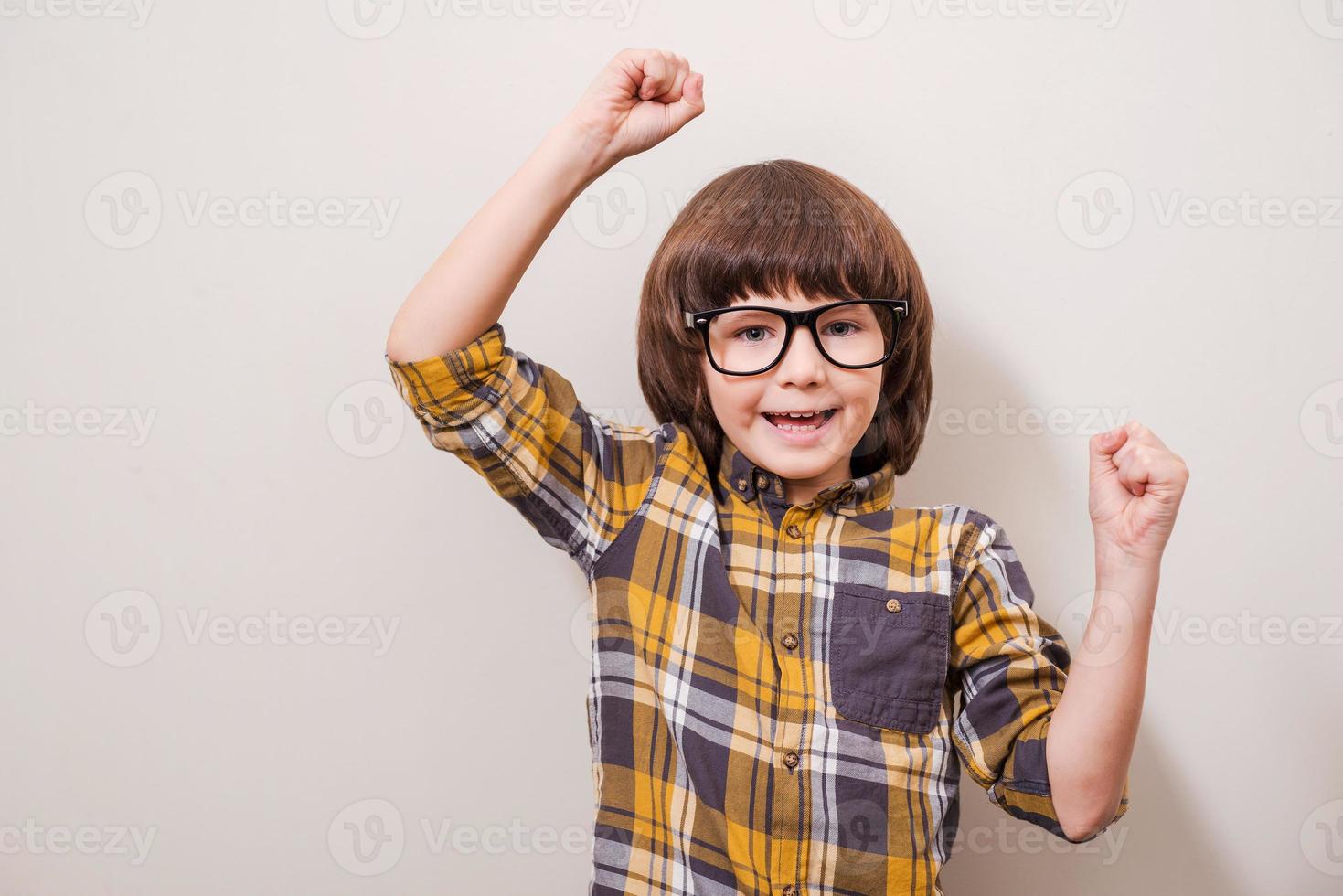 Little things can make him happy Little boy in eyewear keeping arms raised and smiling while standing against grey background photo