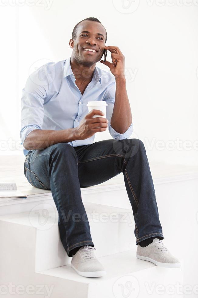buena plática. alegre joven africano con camisa azul sosteniendo una taza de café y hablando por teléfono móvil foto