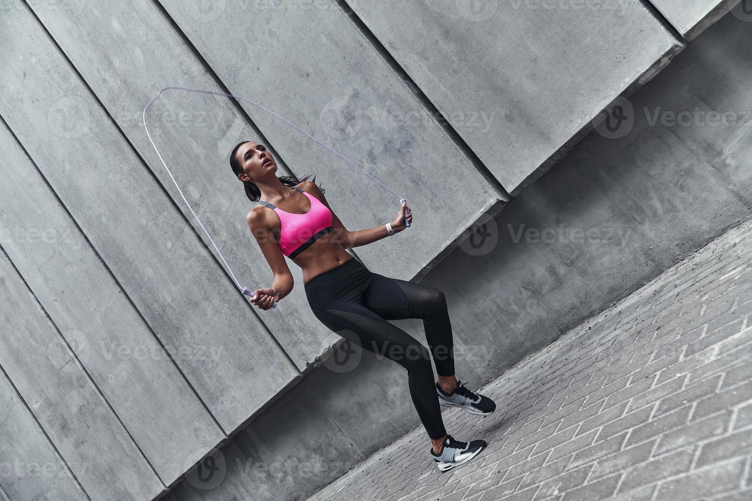 Great shape. Full length of modern young woman in sports clothing skipping rope while exercising outdoors photo