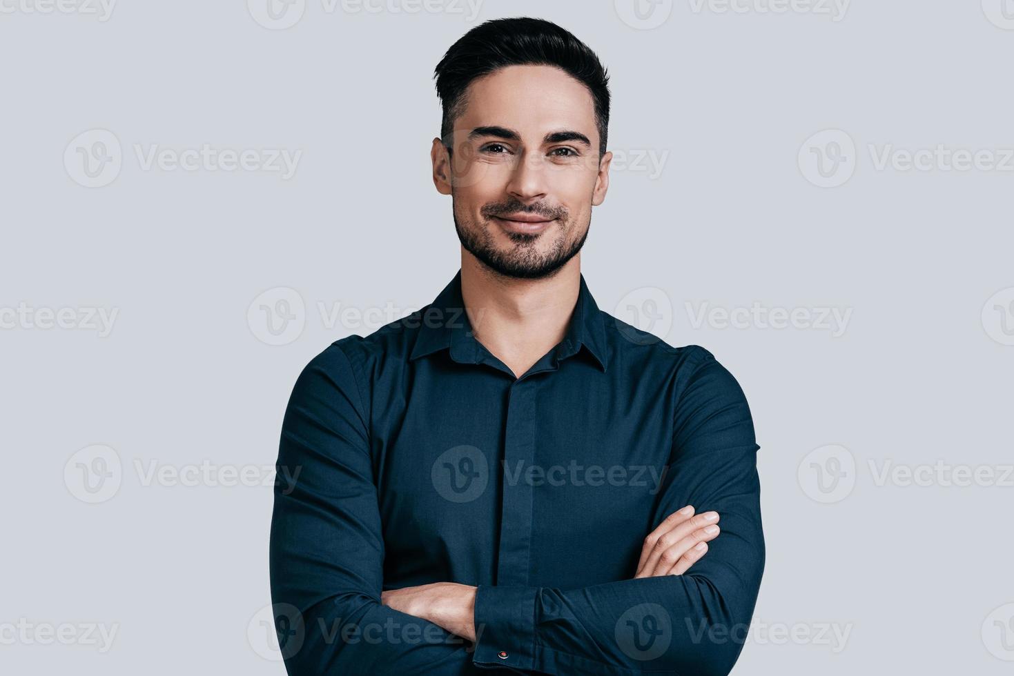 Young and successful. Handsome young man in shirt keeping arms crossed and smiling while standing against grey background photo