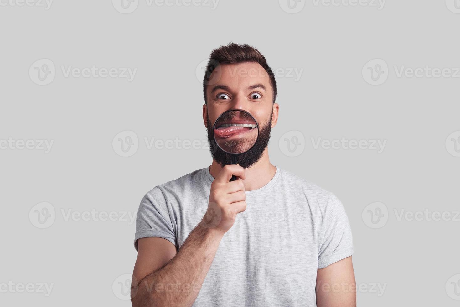 Bizarre man. Playful young man applying magnifying glass and sticking out tongue while standing against grey background photo