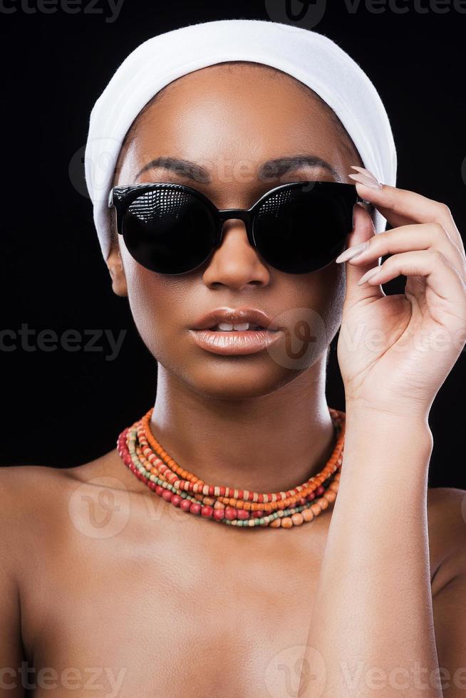 Beauty in style. Beautiful African woman wearing a headscarf and adjusting her sunglasses while standing against black background photo