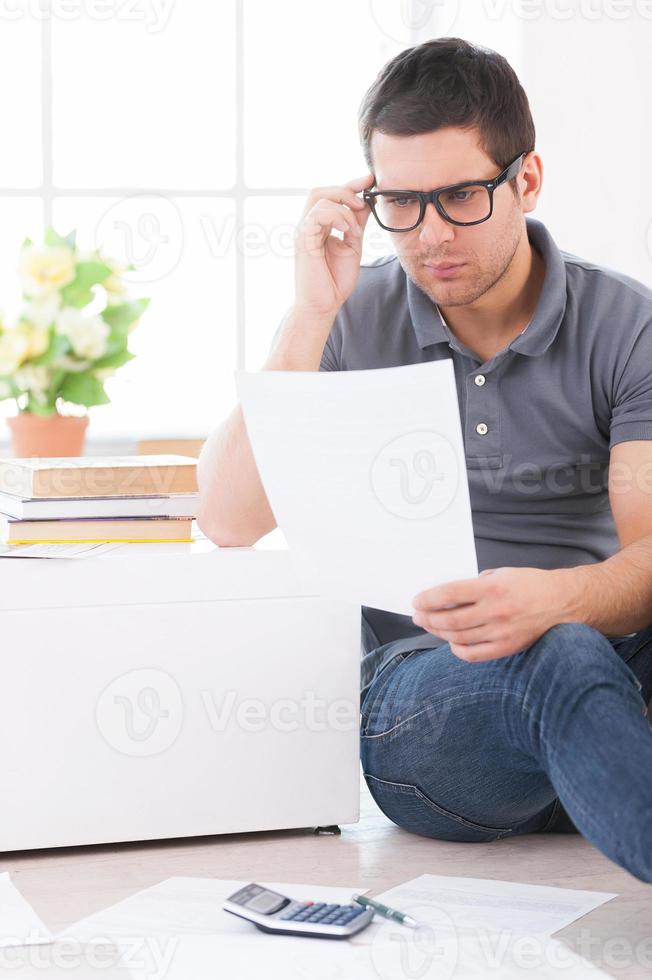 calculando sus gastos. joven concentrado sosteniendo un papel y ajustándose las gafas mientras se sienta en el suelo en su apartamento foto