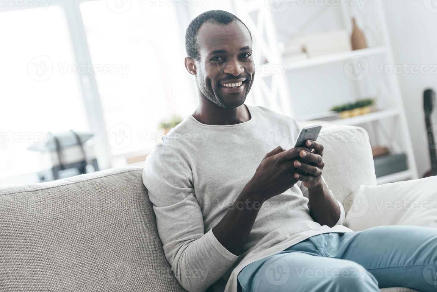 Lazy day at home. Handsome young African man typing a message and looking at camera while sitting on the sofa photo