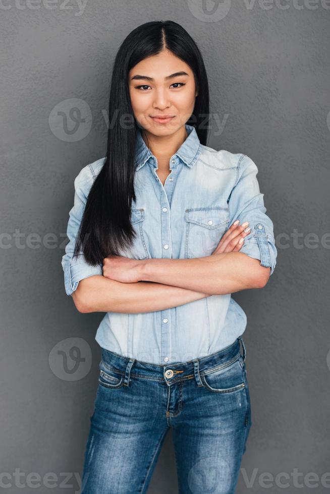 Stylish smile. Beautiful young Asian woman keeping arms crossed and looking at camera with smile while standing against grey background photo