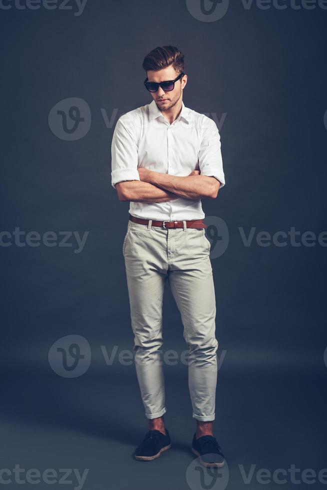 Casually handsome. Full length of confident young handsome man in sunglasses keeping arms crossed and looking down while standing against grey background photo