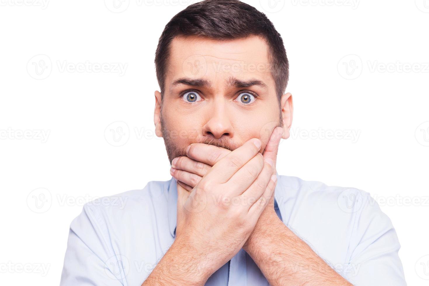 No way Shocked young man in formalwear covering mouth with hands and looking at camera while standing against white background photo