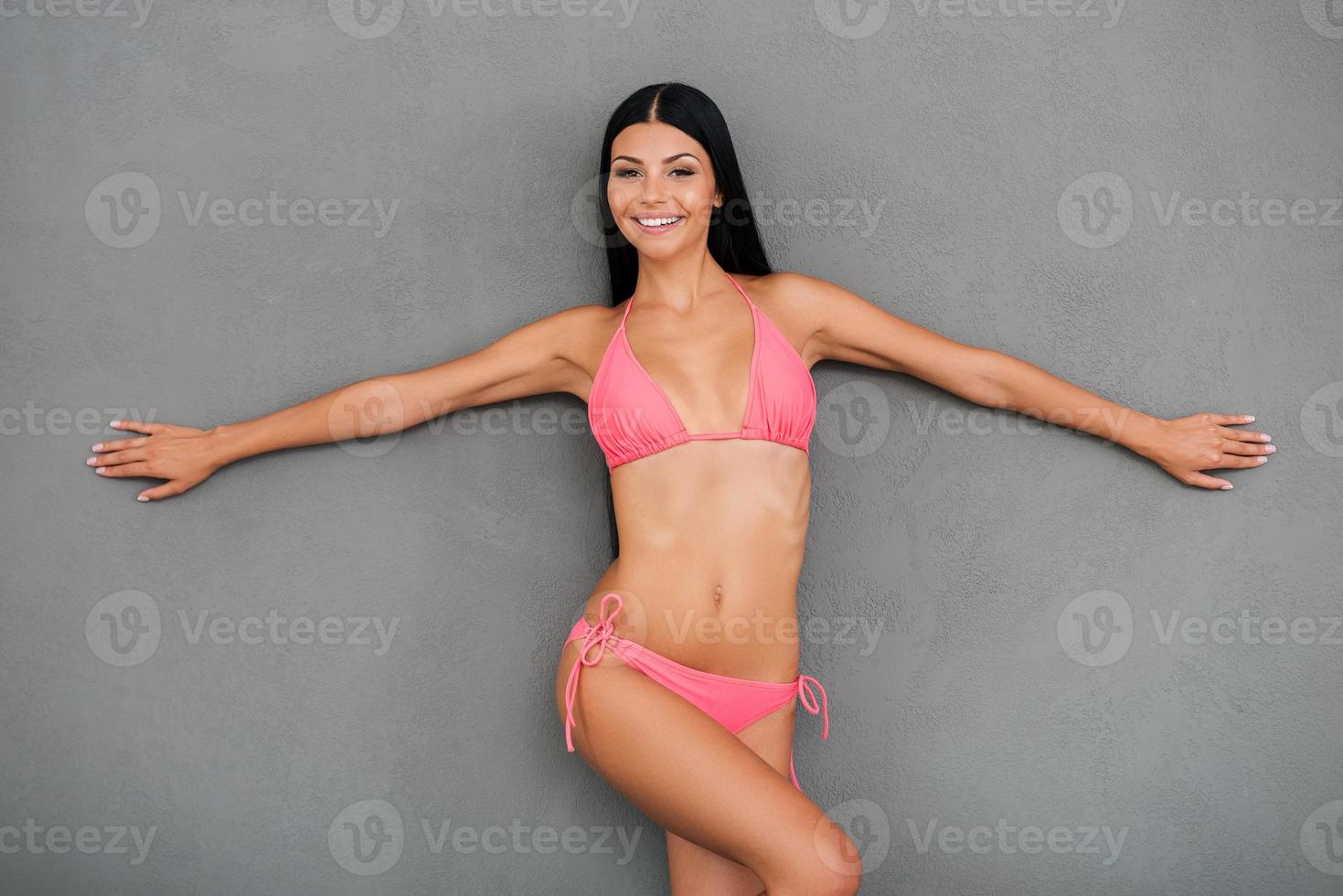 Ready for summer Happy young woman in swimsuit keeping arms outstretched and smiling while posing against grey background photo
