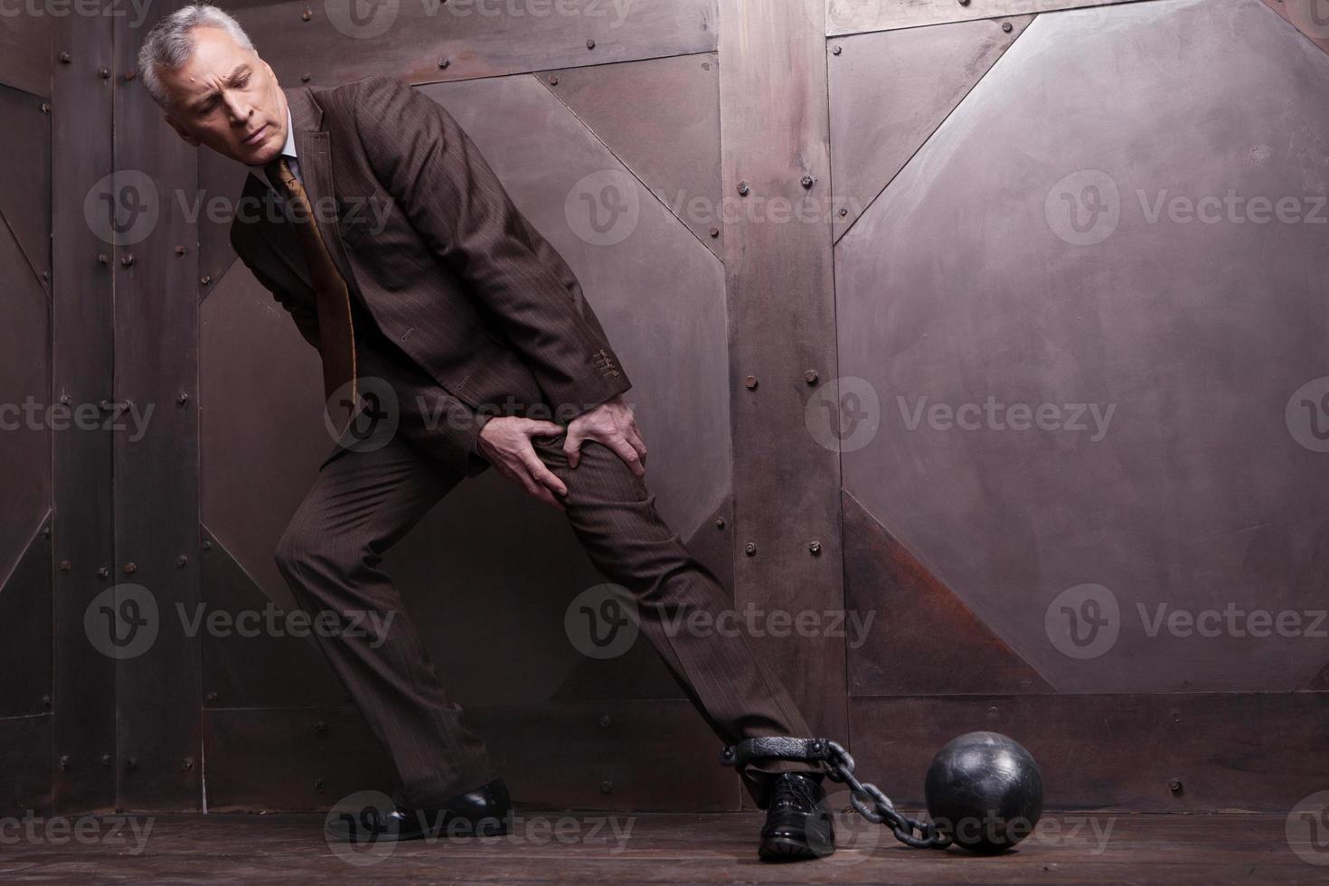 Chains on his leg. Full length of grey hair senior man in formalwear trying to go with the shackles on his leg photo