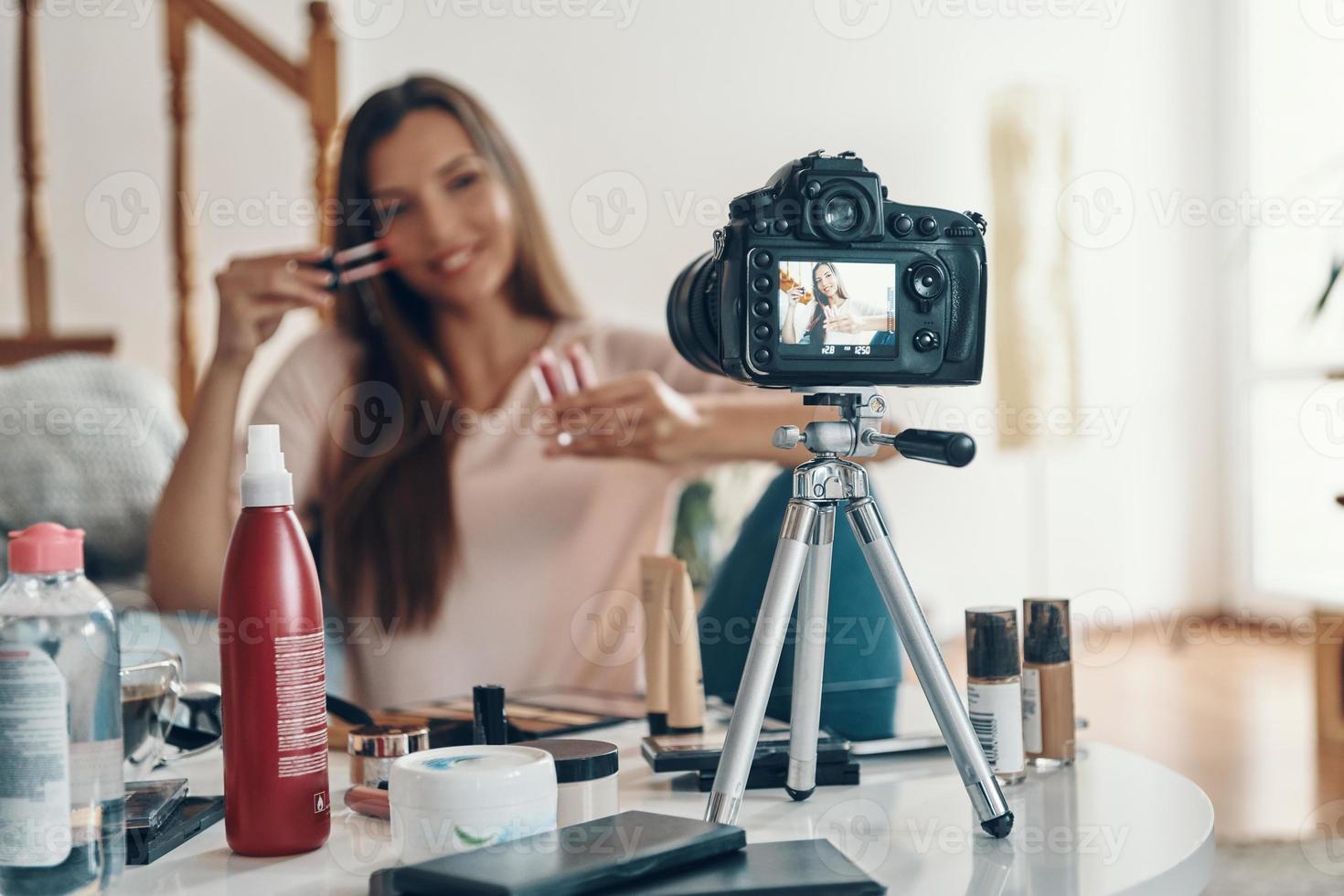 Good looking young woman applying lip gloss and smiling while making social media video photo