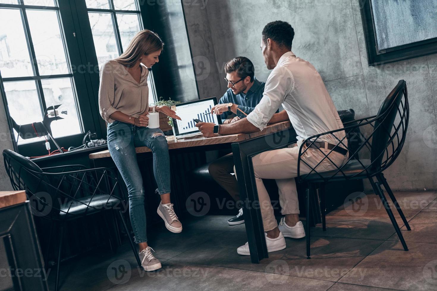 Three young people in smart casual wear analyzing data on laptop while working in the office photo
