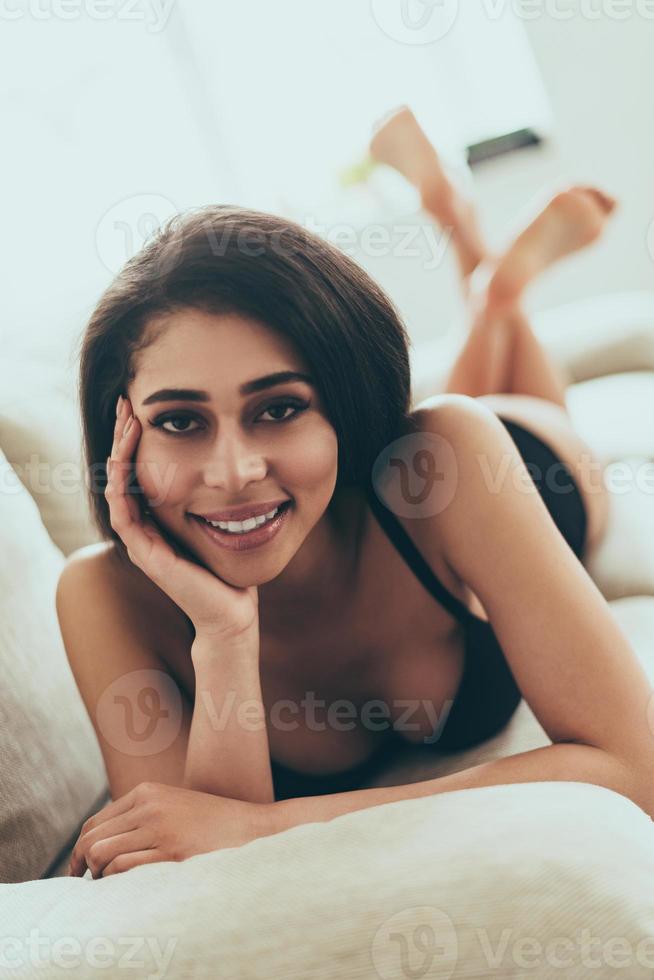 belleza coqueta. hermosa joven mujer de raza mixta en lencería negra mirando a la cámara y sonriendo mientras está acostada en el sofá en casa foto