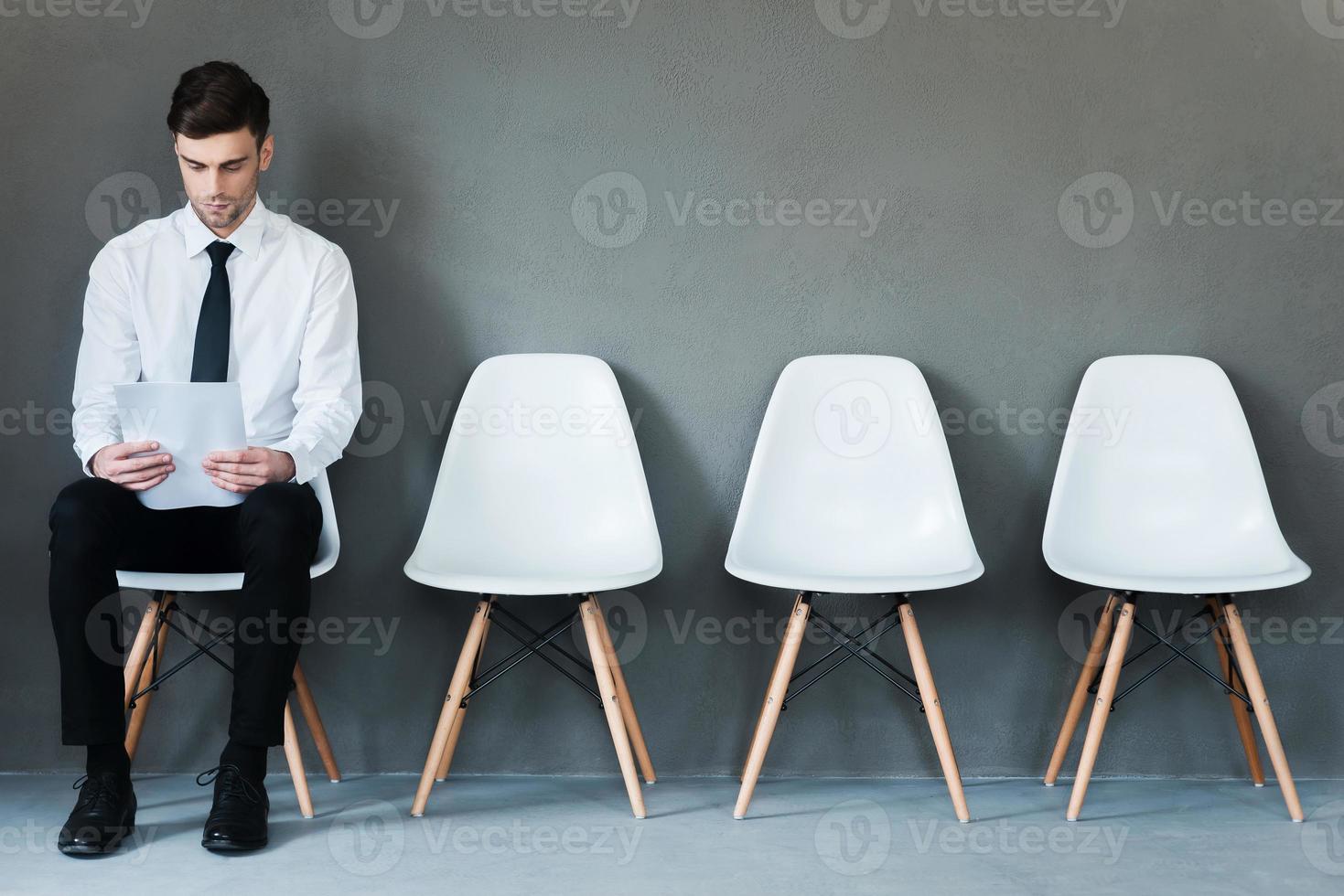 Waiting for interview. Confident young businessman holding paper while sitting on chair against grey background photo