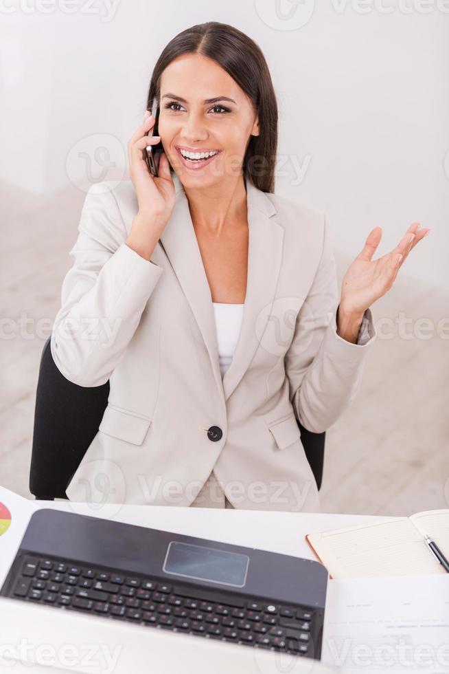 Good business talk. Top view of beautiful young businesswoman talking on the mobile phone and smiling while sitting at her working place photo
