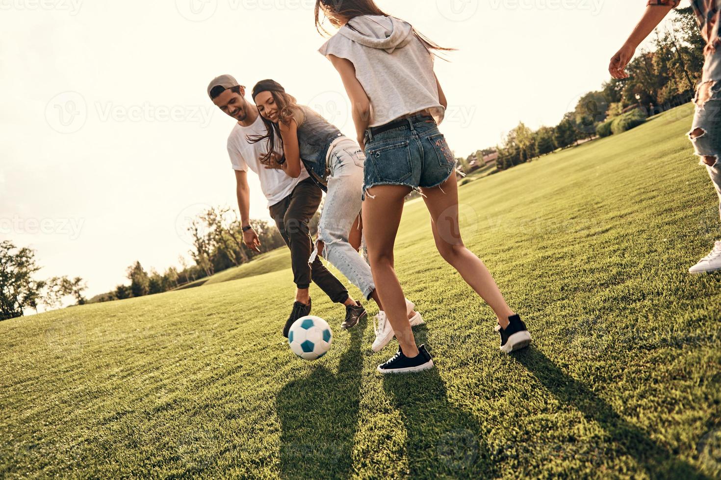 Trying to win. Group of young smiling people in casual wear enjoying nice summer day while playing soccer outdoors photo