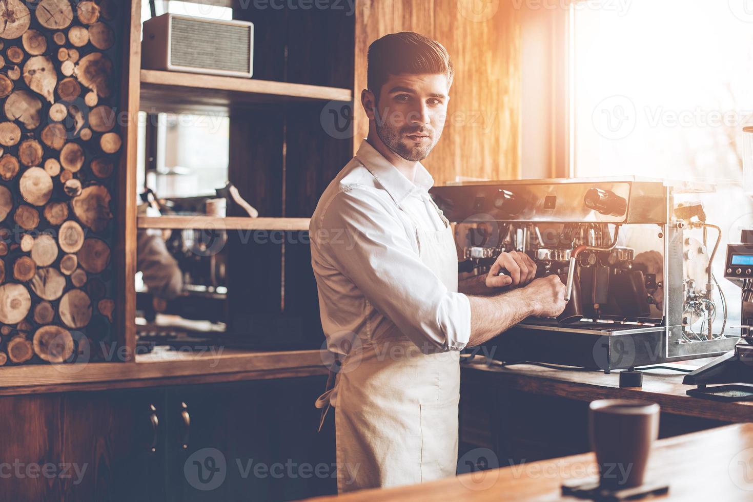Loving his job. Young man in apron making coffee and looking at camera while standing at cafe photo