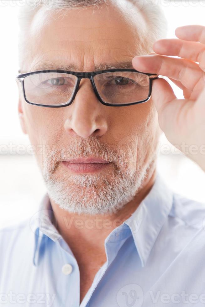 Confident look. Confident grey hair senior man in shirt adjusting his eyeglasses and looking at camera while standing in front of the window photo