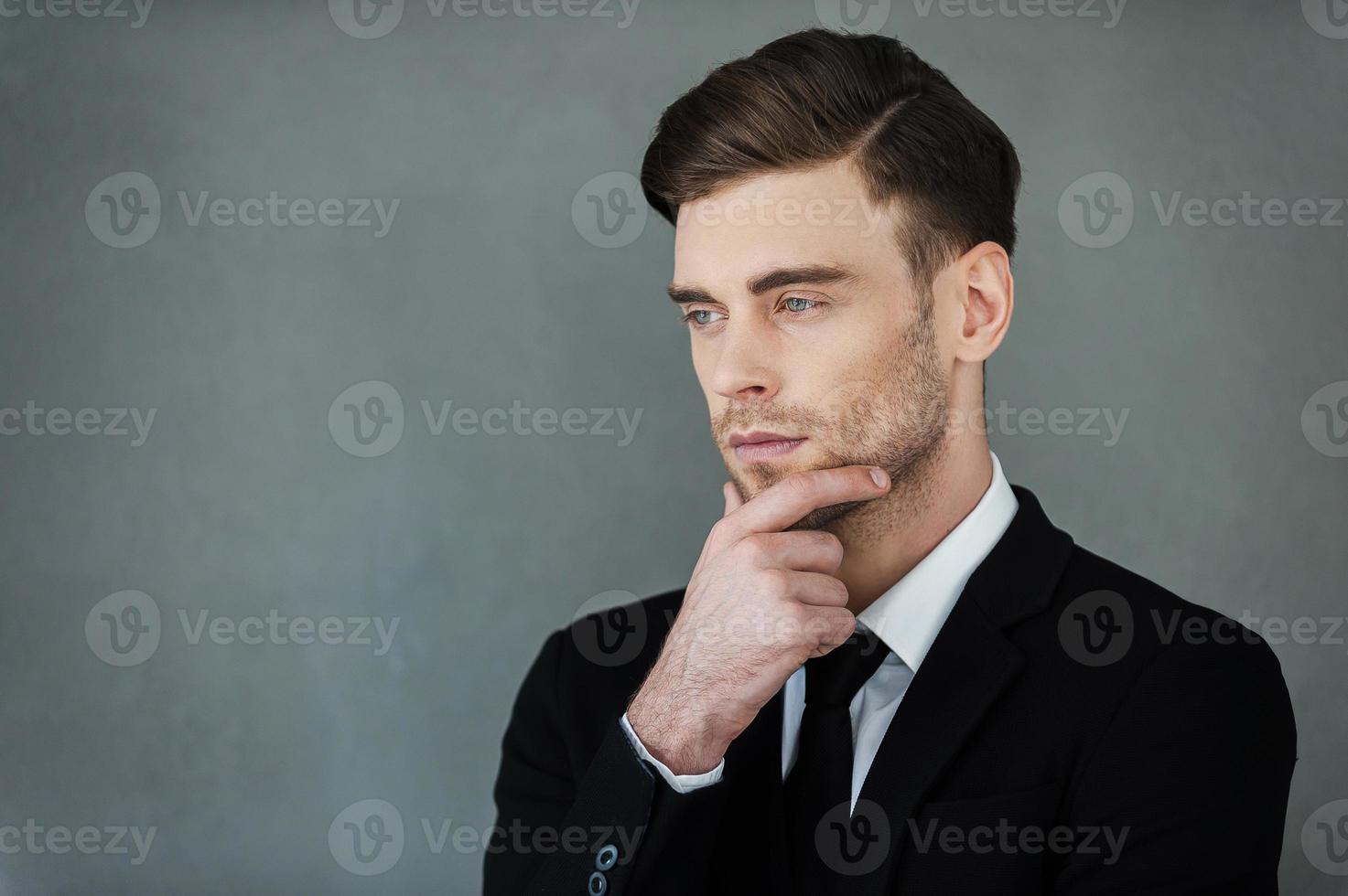 Elegance and confidence. Thoughtful young businessman holding hand on chin and looking away while standing against grey background photo