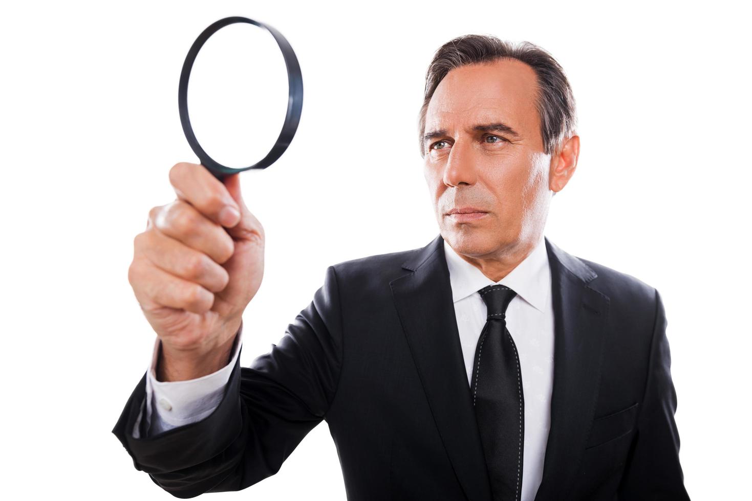 Businessman with magnifying glass. Concentrated mature man in formalwear looking through a magnifying glass while standing isolated on white background photo