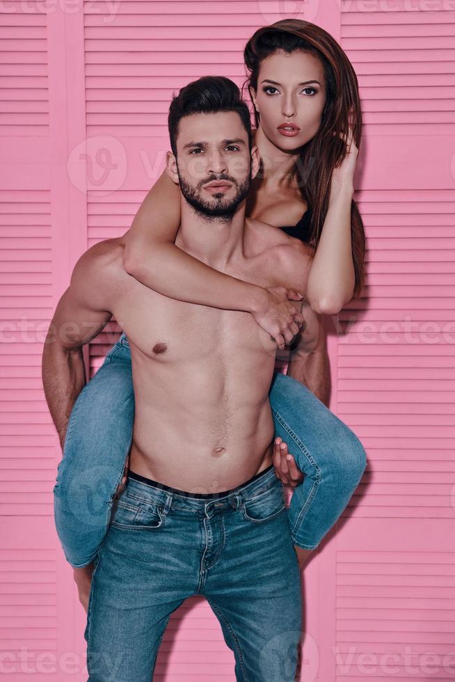 Feeling loved. Handsome shirtless man carrying his girlfriend on shoulders while standing against pink background photo