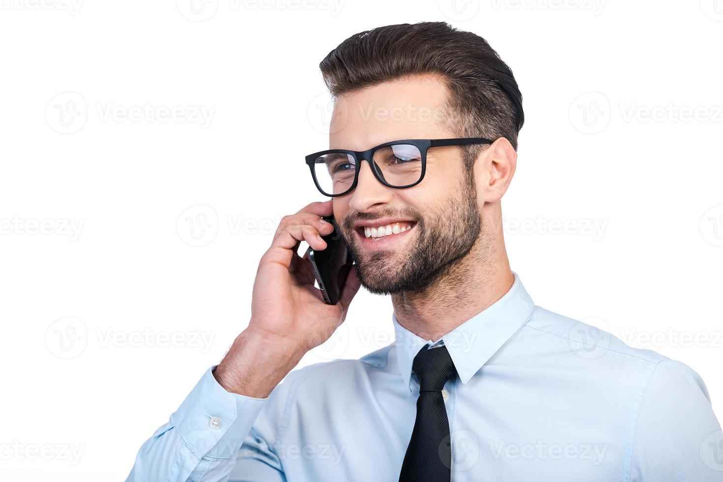 buena charla de negocios. hombre guapo joven confiado en camisa y corbata hablando por teléfono móvil y sonriendo mientras está de pie contra el fondo blanco foto