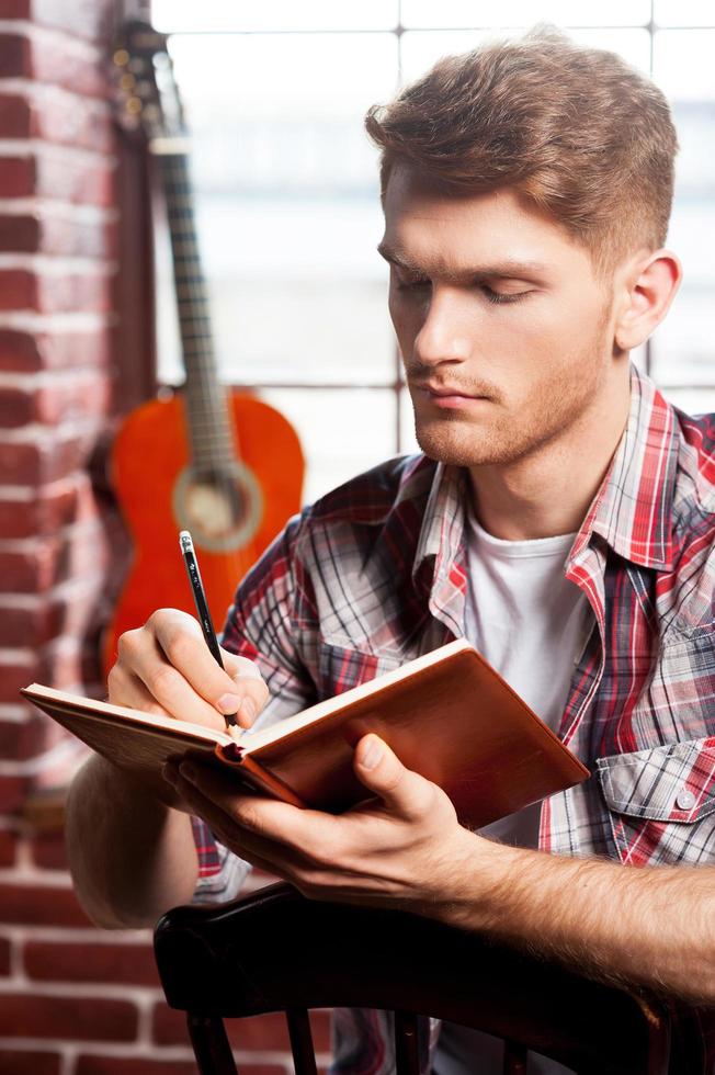 compositor creativo. un joven apuesto escribiendo algo en un bloc de notas mientras la guitarra acústica está en el fondo foto