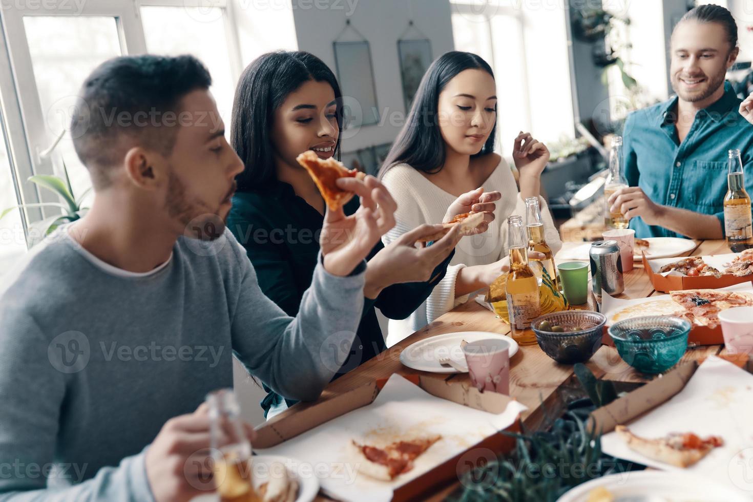 Dinner with friends. Group of young people in casual wear eating pizza and smiling while having a dinner party indoors photo