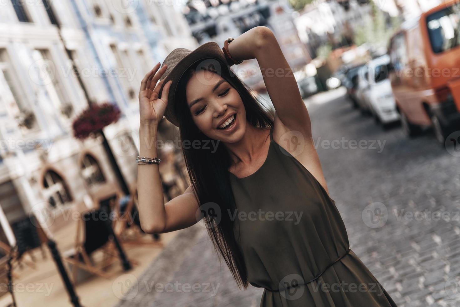 chica de moda en la ciudad. mujer joven atractiva ajustando su sombrero y sonriendo mientras está de pie al aire libre foto