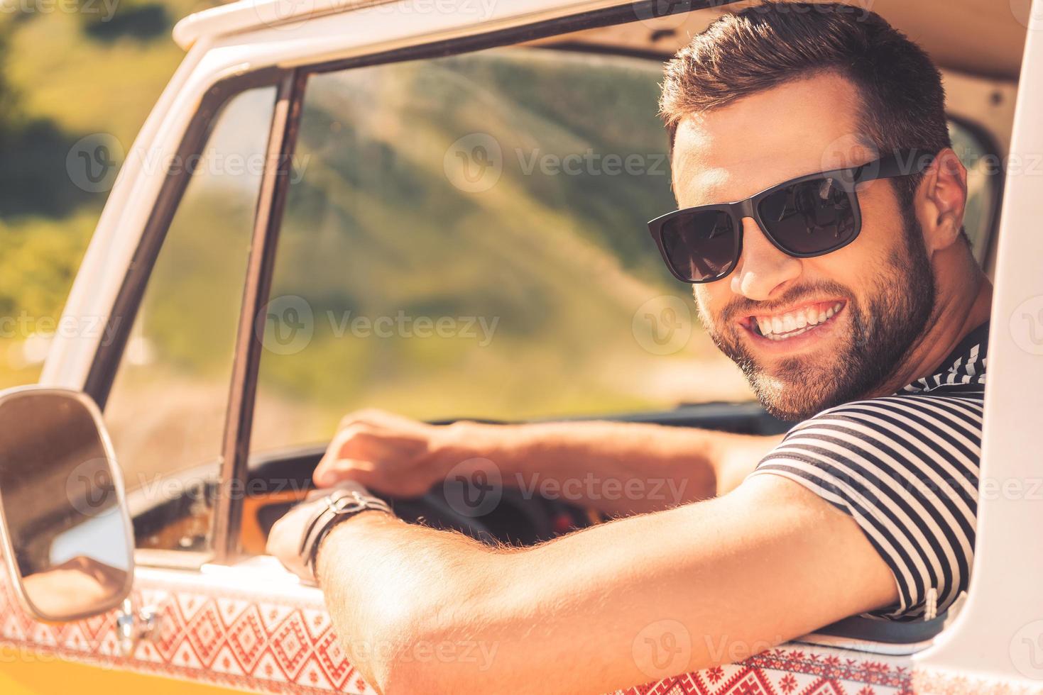 Enjoying his road trip. Cheerful young man smiling at camera and holding hand on steering wheel while sitting inside of his minivan photo