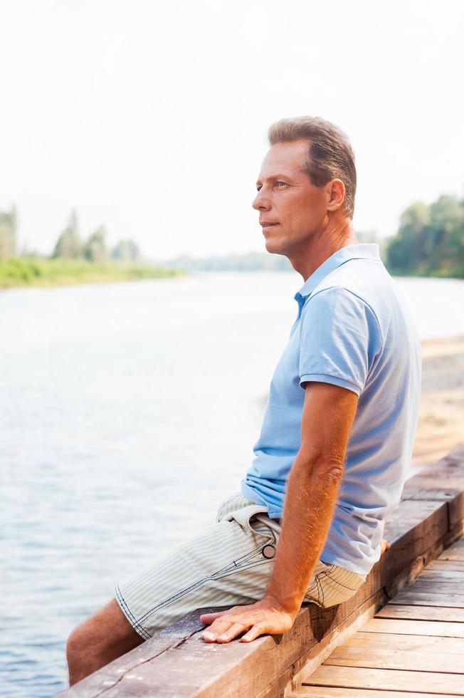 Day dreaming on riverbank. Side view of thoughtful mature man looking away while sitting at the quayside photo