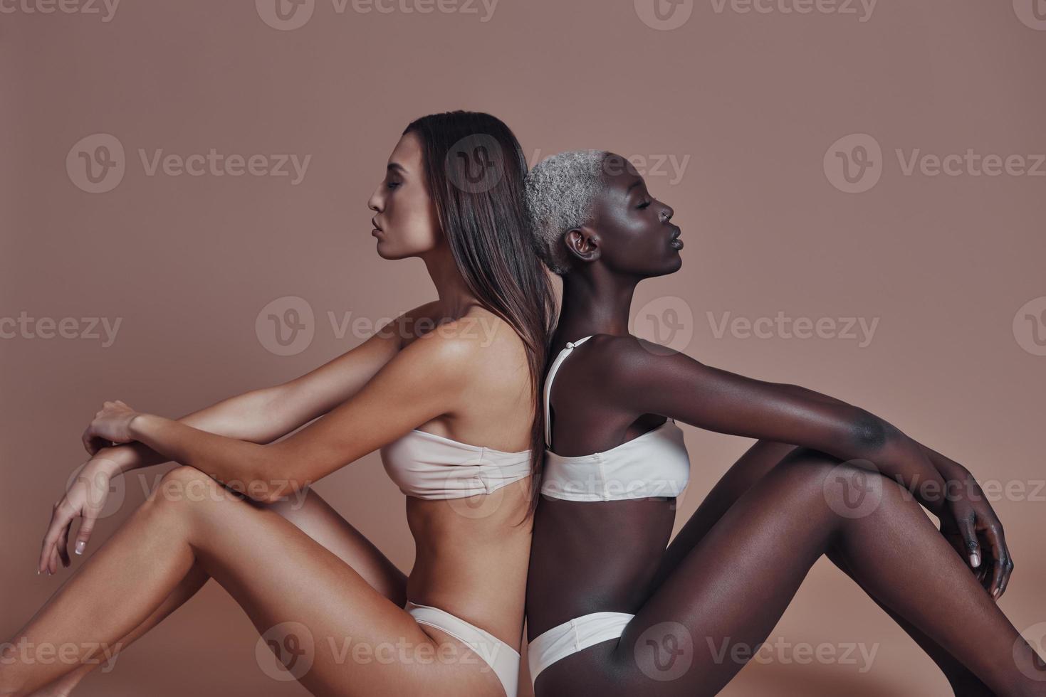 True beauty. Two attractive mixed race women looking away while sitting back to back against brown background photo