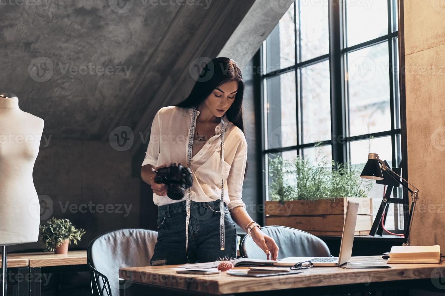 Developing new fashion line. Concentrated young woman with tape measure on her neck looking at fashion magazine and holding digital camera while standing near the desk in her workshop photo