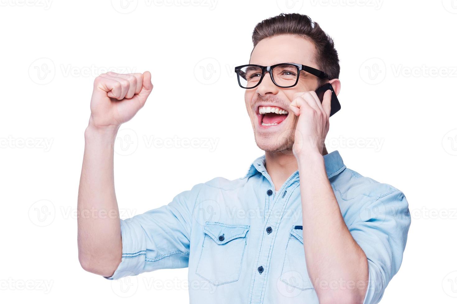 Great news Handsome young man in shirt holding mobile phone and gesturing while standing against white background photo