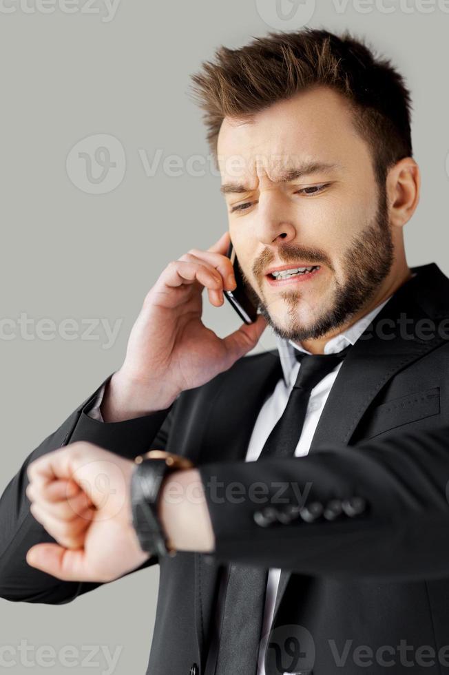 I will be in time Portrait of worried young man in formalwear talking on the phone and checking the time while standing against grey background photo