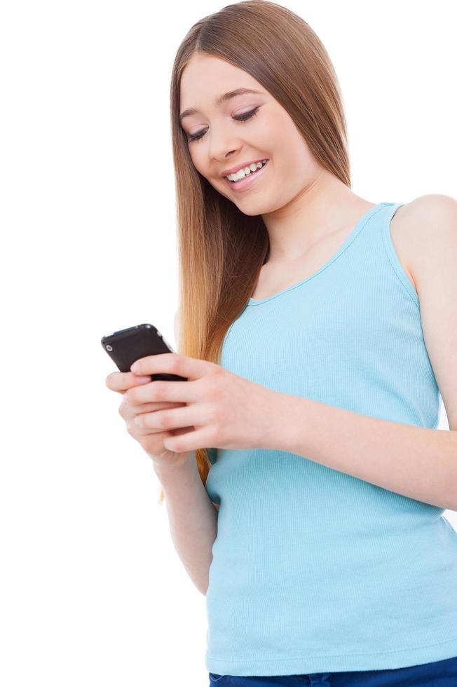 Teenage typing message. Cheerful teenage girl holding mobile phone and looking at it while standing isolated on white photo