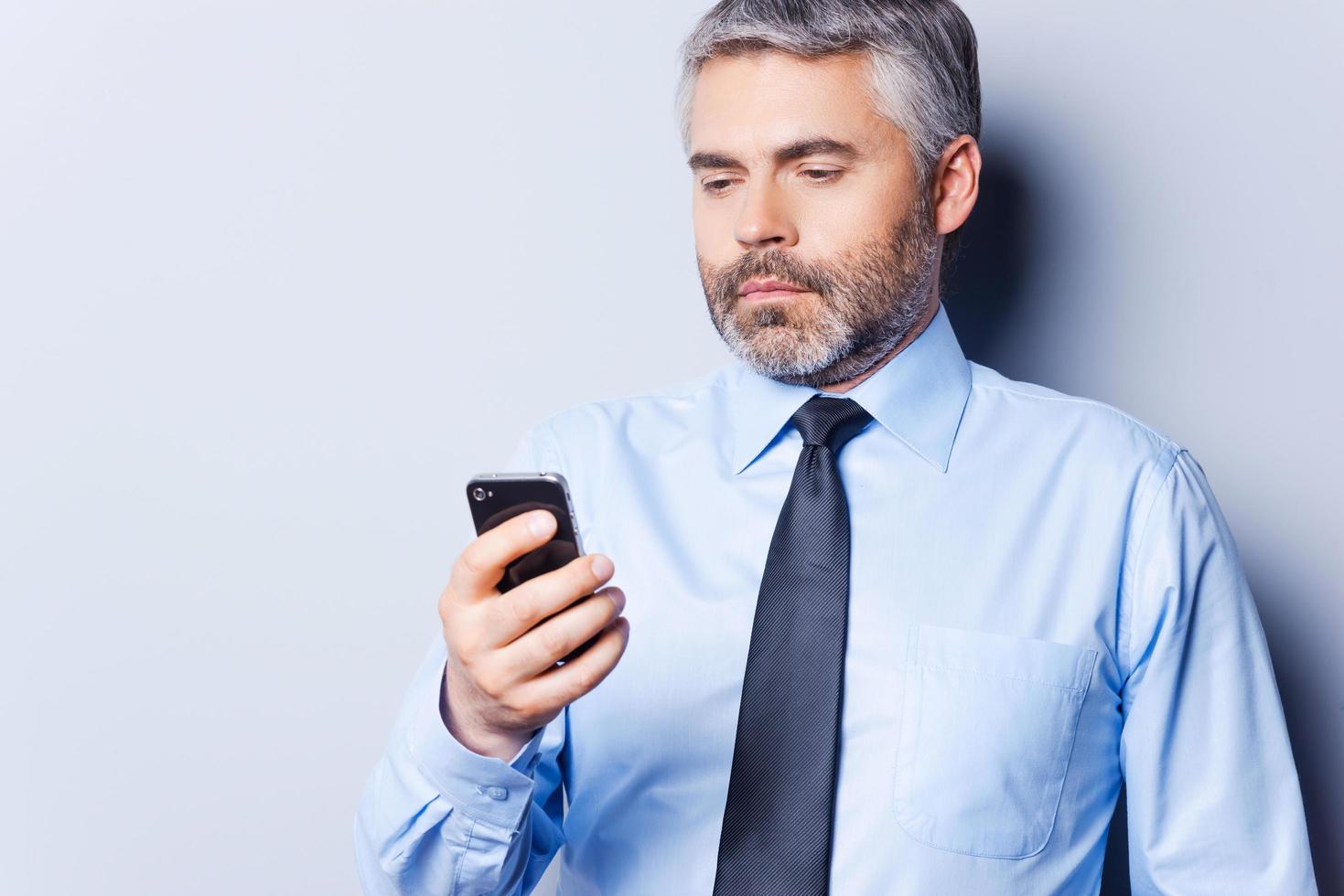 revisando mensajes de negocios. hombre maduro seguro de sí mismo con camisa y corbata sosteniendo un teléfono móvil y mirándolo mientras se enfrenta a un fondo gris foto