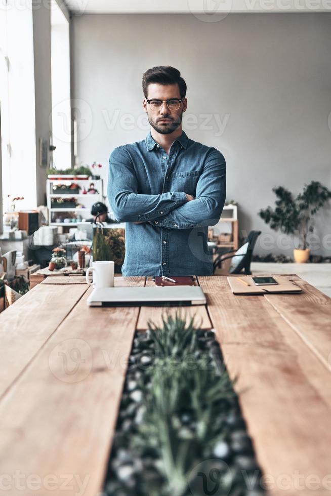 Young and successful. Handsome young man looking a camera and keeping arms crossed while standing in the creative working space photo