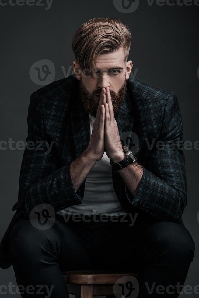 Perfect man. Beautiful young man keeping hands clasped and looking at camera while sitting against grey background photo