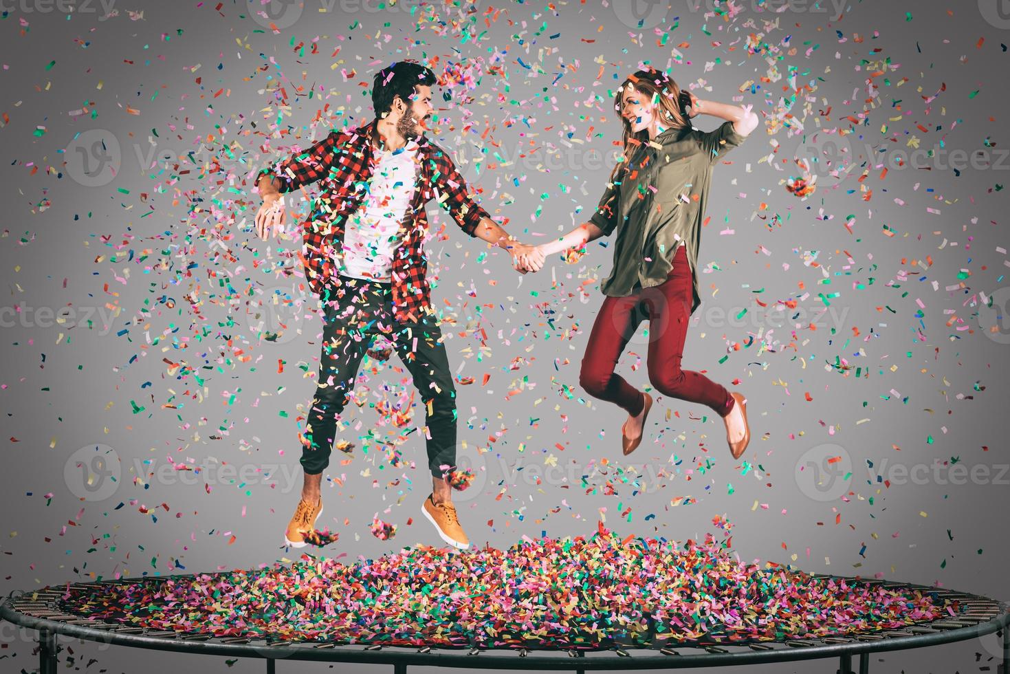 Carefree fun. Mid-air shot of beautiful young cheerful couple holding hands while jumping on trampoline together with confetti all around them photo