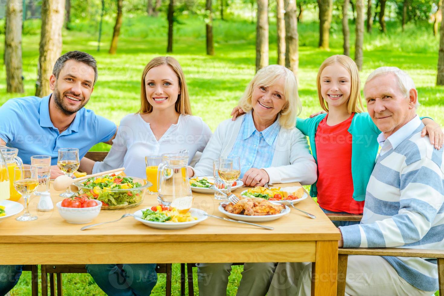 familia junta. familia feliz de cinco personas que se unen y sonríen mientras se sientan en la mesa del comedor al aire libre foto