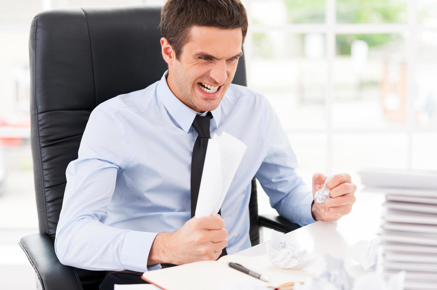 Sick and tired. Furious young man in shirt and tie holding papers in hands and shouting while sitting at his working place photo