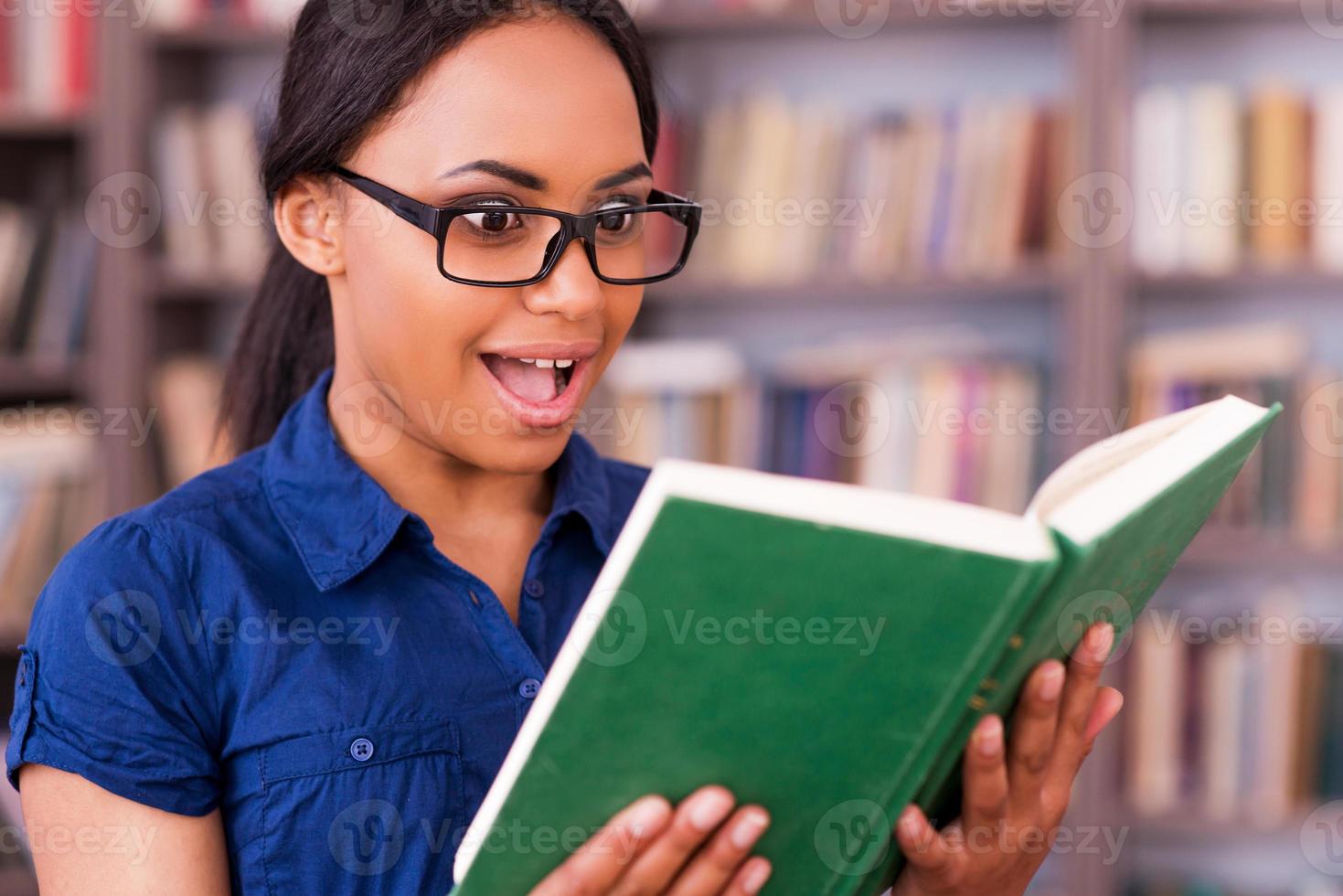 What an exciting book Excited African female student reading a book and keeping mouth open while standing in library photo