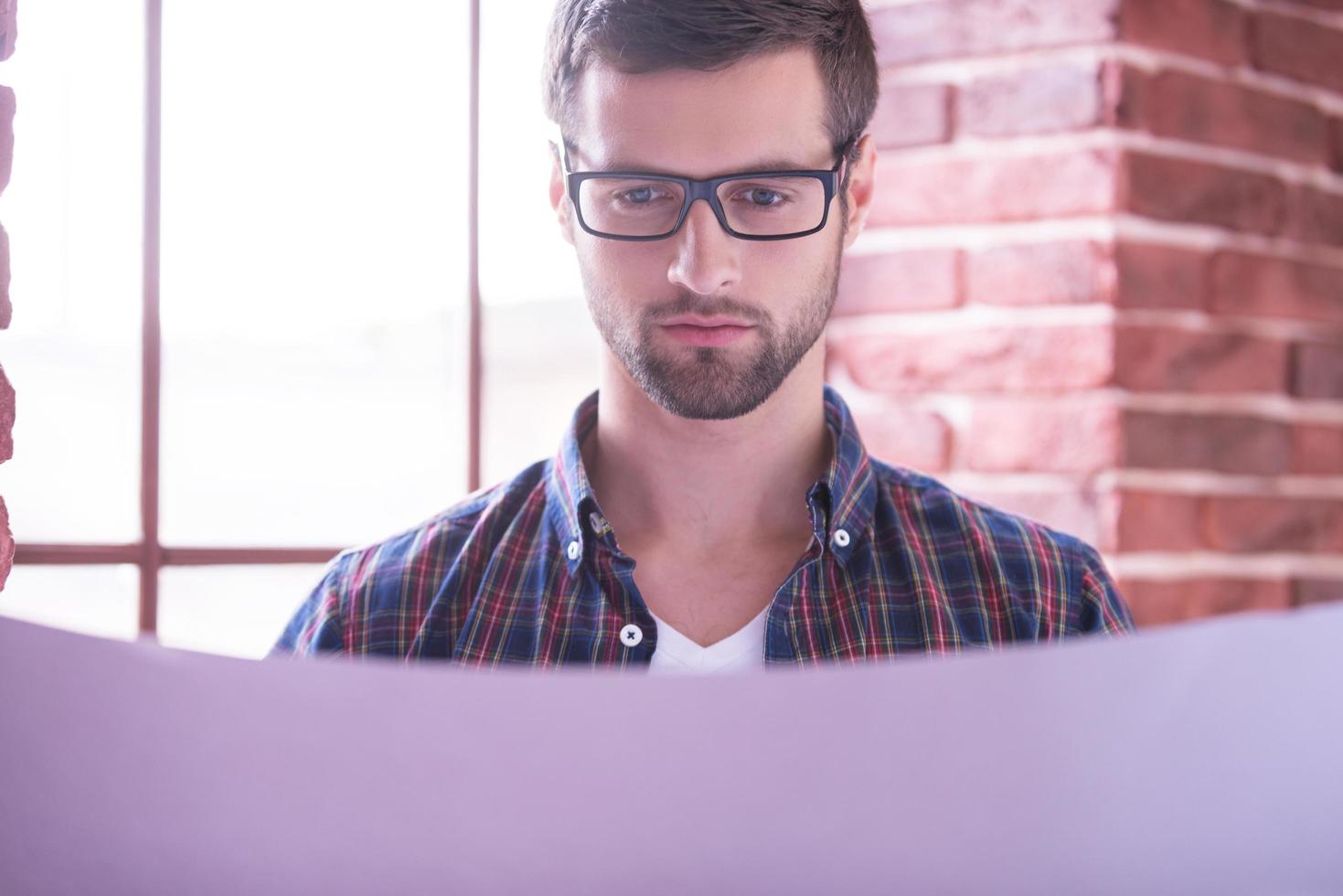 Young and confident architect. Handsome young man in glasses examining blueprint while standing near the window photo