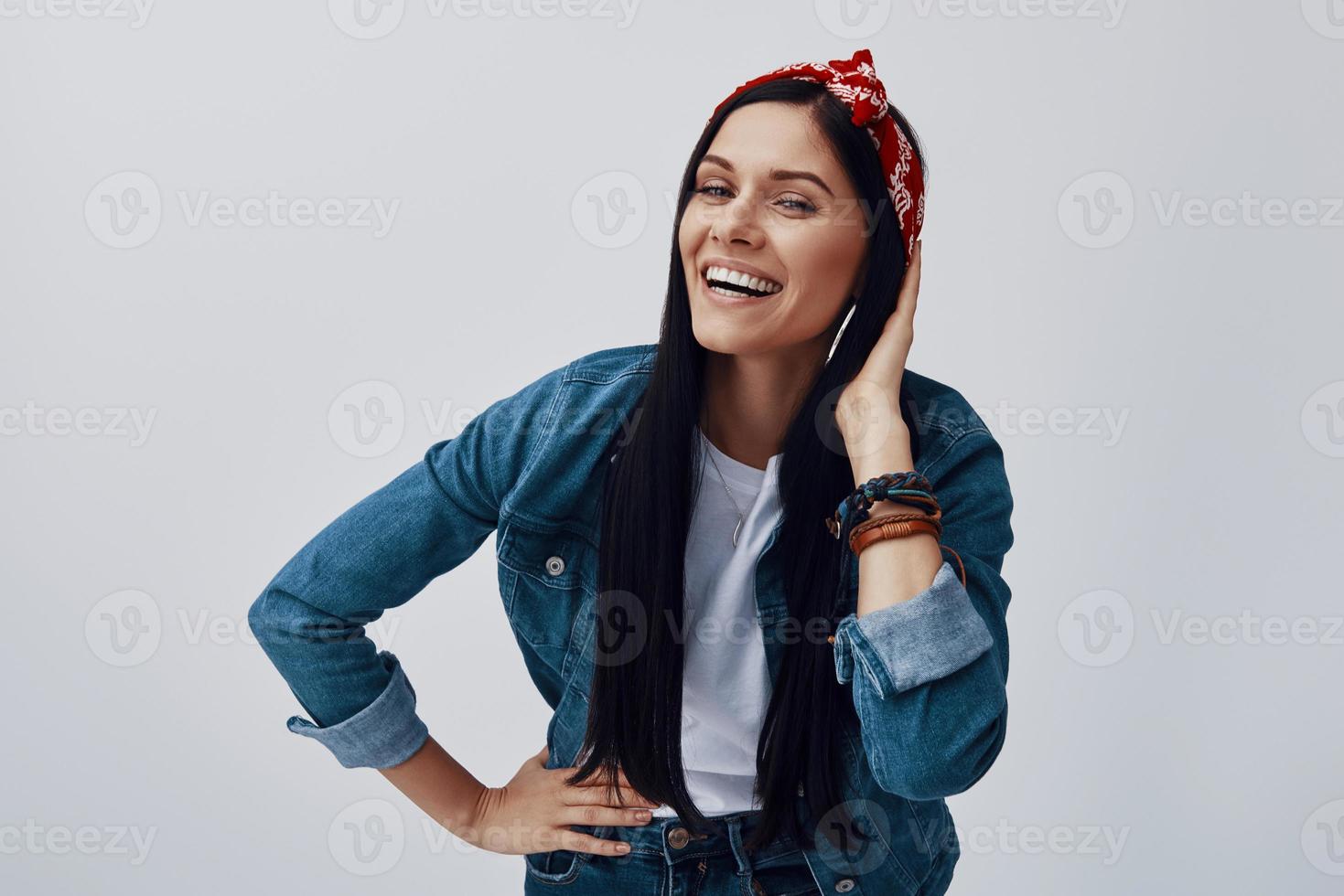 Funky young woman in bandana looking at camera and smiling while standing against grey background photo