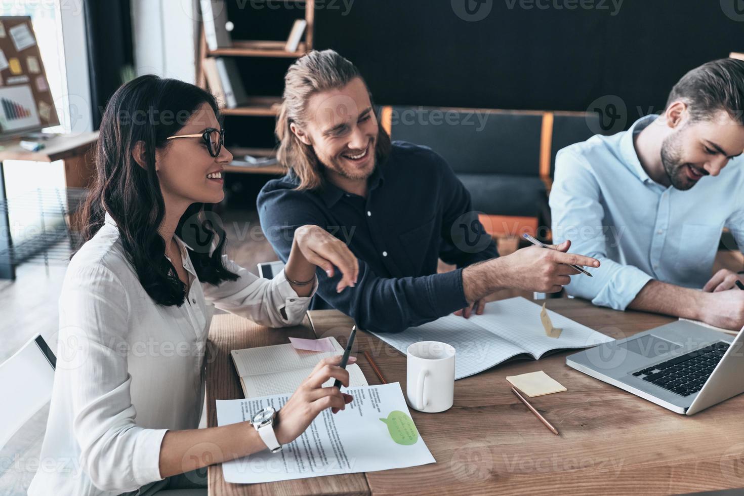 Discussing risky ideas. Group of young modern people in smart casual wear pointing at laptop and smiling while sitting in the creative office photo
