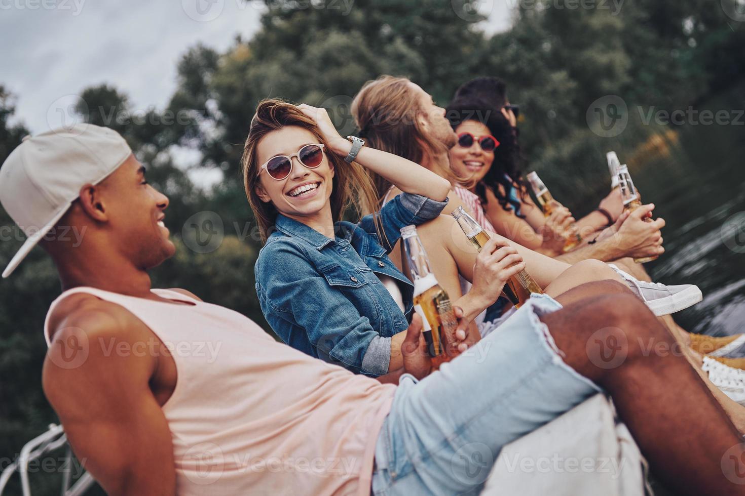 Leave all the worries behind. Group of happy young people in casual wear smiling and drinking beer while sitting on the pier photo