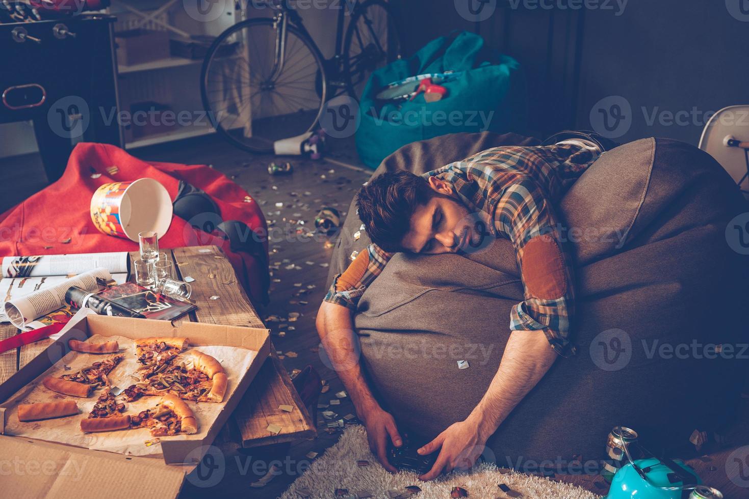 lugar cómodo para desmayarse. joven apuesto se desmayó en una bolsa de frijoles con joystick en la mano en una habitación desordenada después de la fiesta foto