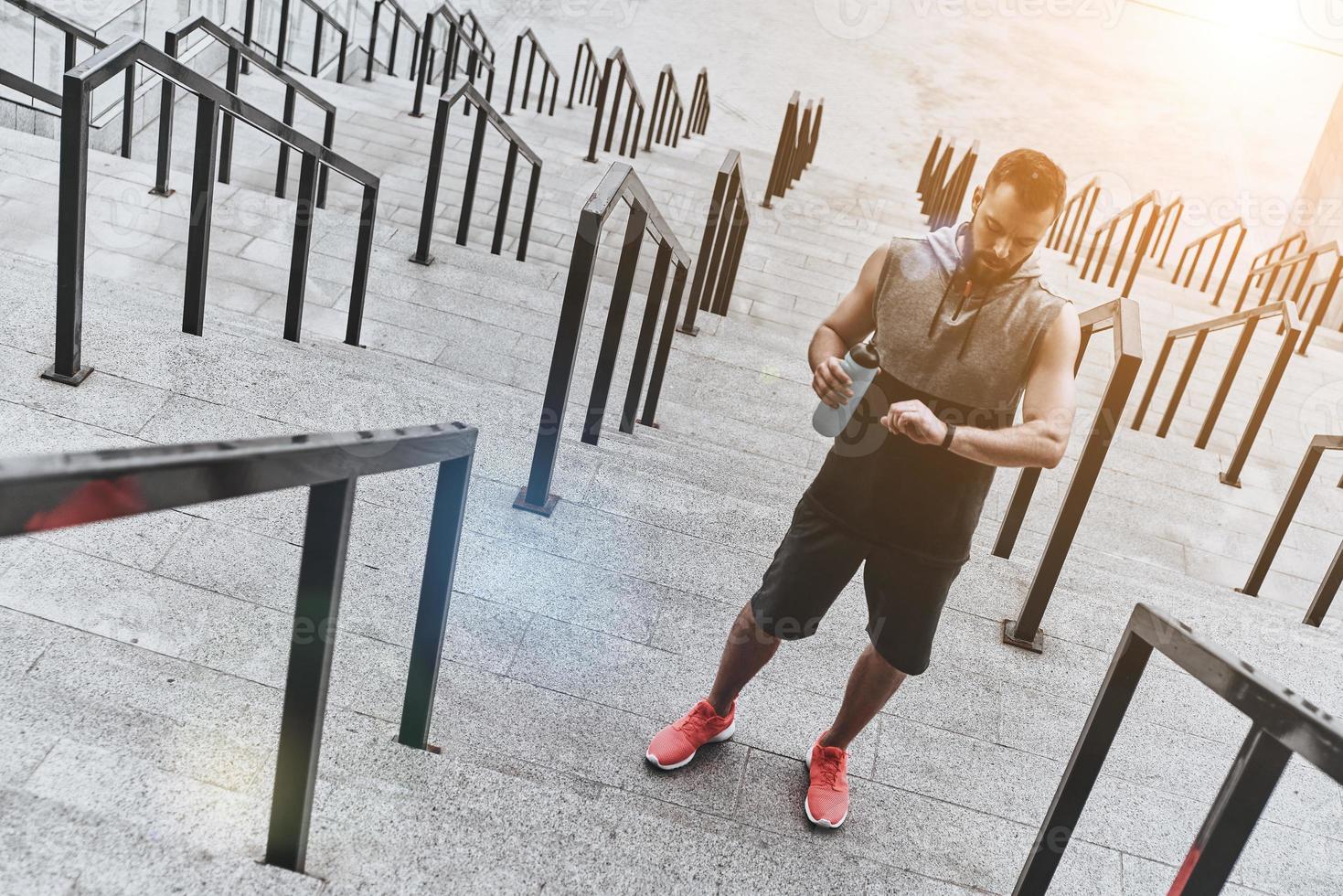Having short rest. Handsome young man in sport clothing looking at his fitness tracker while standing on the steps outside photo
