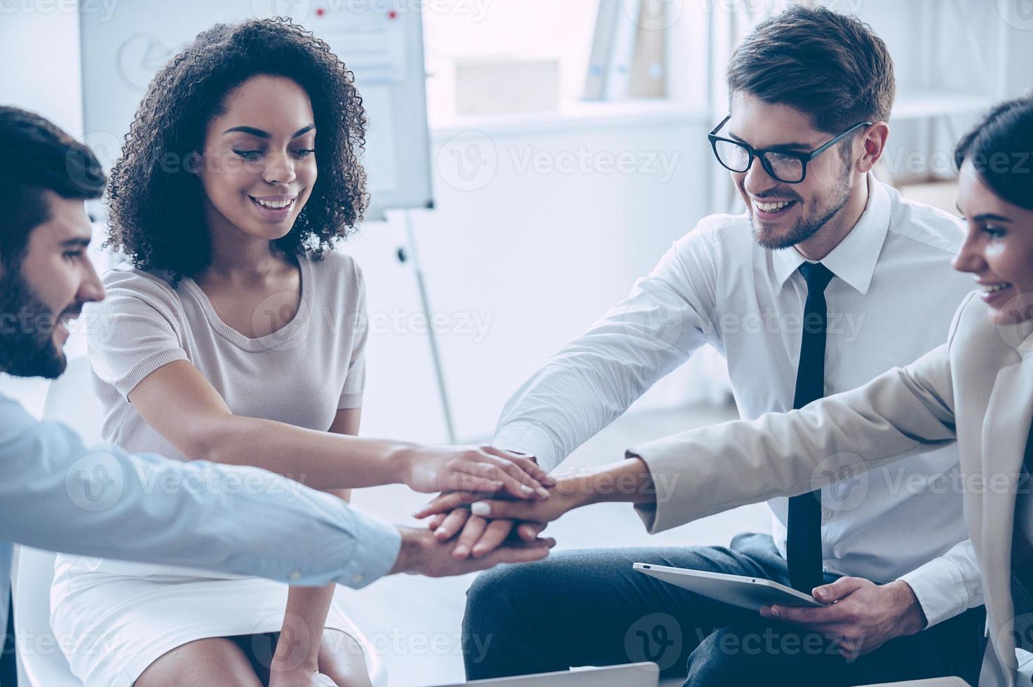 New achievements are welcome  Close-up part of top view of group of four young people holding hands and showing their unity with smile while sitting at office photo