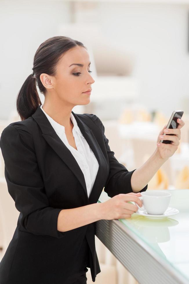 Checking business messages. Confident young woman in formalwear drinking coffee and looking at her mobile phone while leaning at bar counter photo